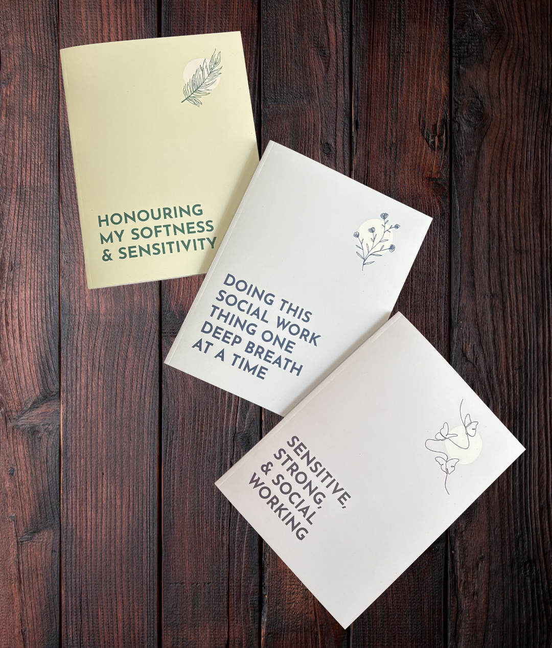 All 3 Sensitive Social Worker Notebooks laid out on a wooden surface.
The Purple Notebook features 3 butterflies and reads "Sensitive, Strong & Social Working".
The Blue Notebook features a plant with 5 blossoms and states "Doing This Social Work Thing One Deep Breath At A Time".
The Green Notebook includes a sketch of a feather and the text "Honouring My Softness & Sensitivity"