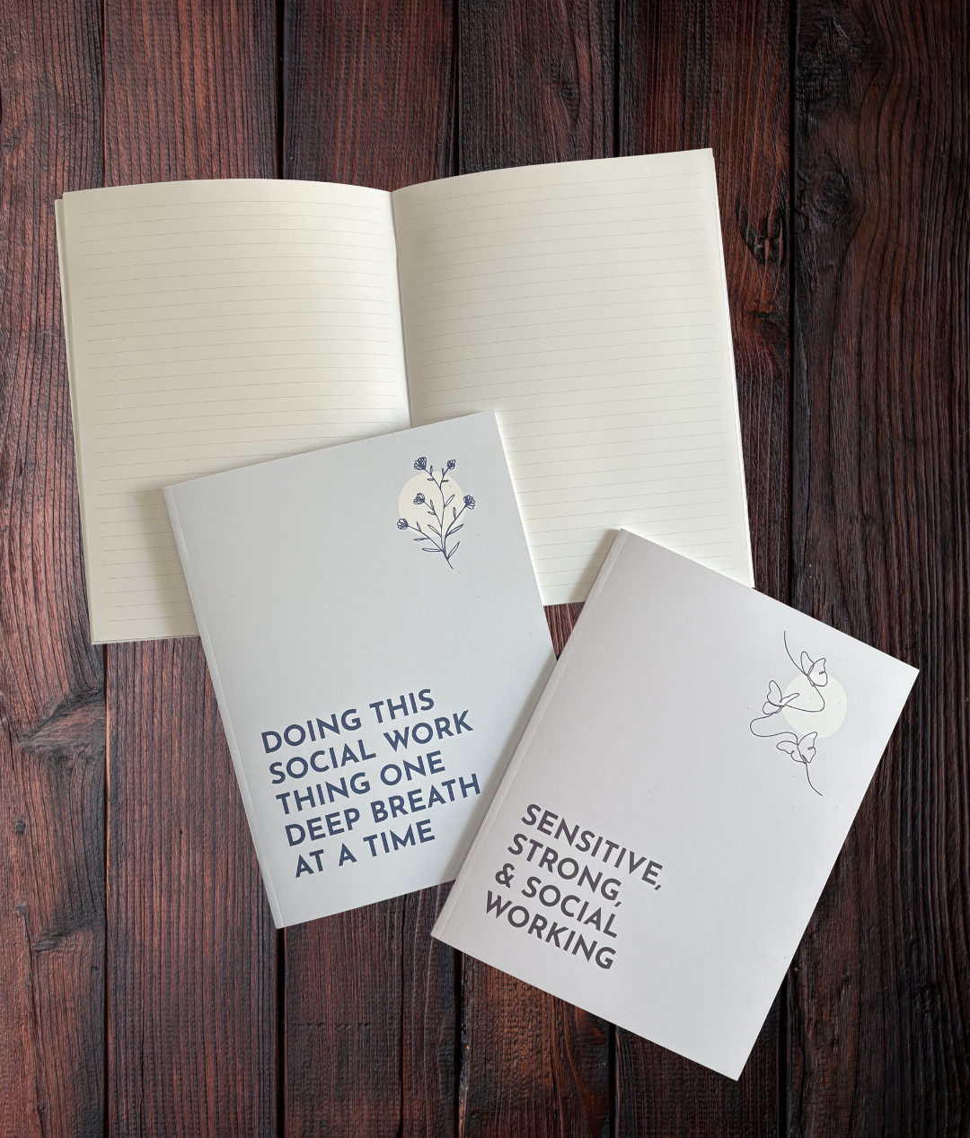 3 Sensitive Social Worker Notebooks on a wooden surface. 1 is open showing lined pages. You can see the covers of the other 2 notebooks: The Purple Notebook which features 3 butterflies and reads "Sensitive, Strong & Social Working" and the Blue Notebook which features a plant with 5 blossoms and states "Doing This Social Work Thing One Deep Breath At A Time".
