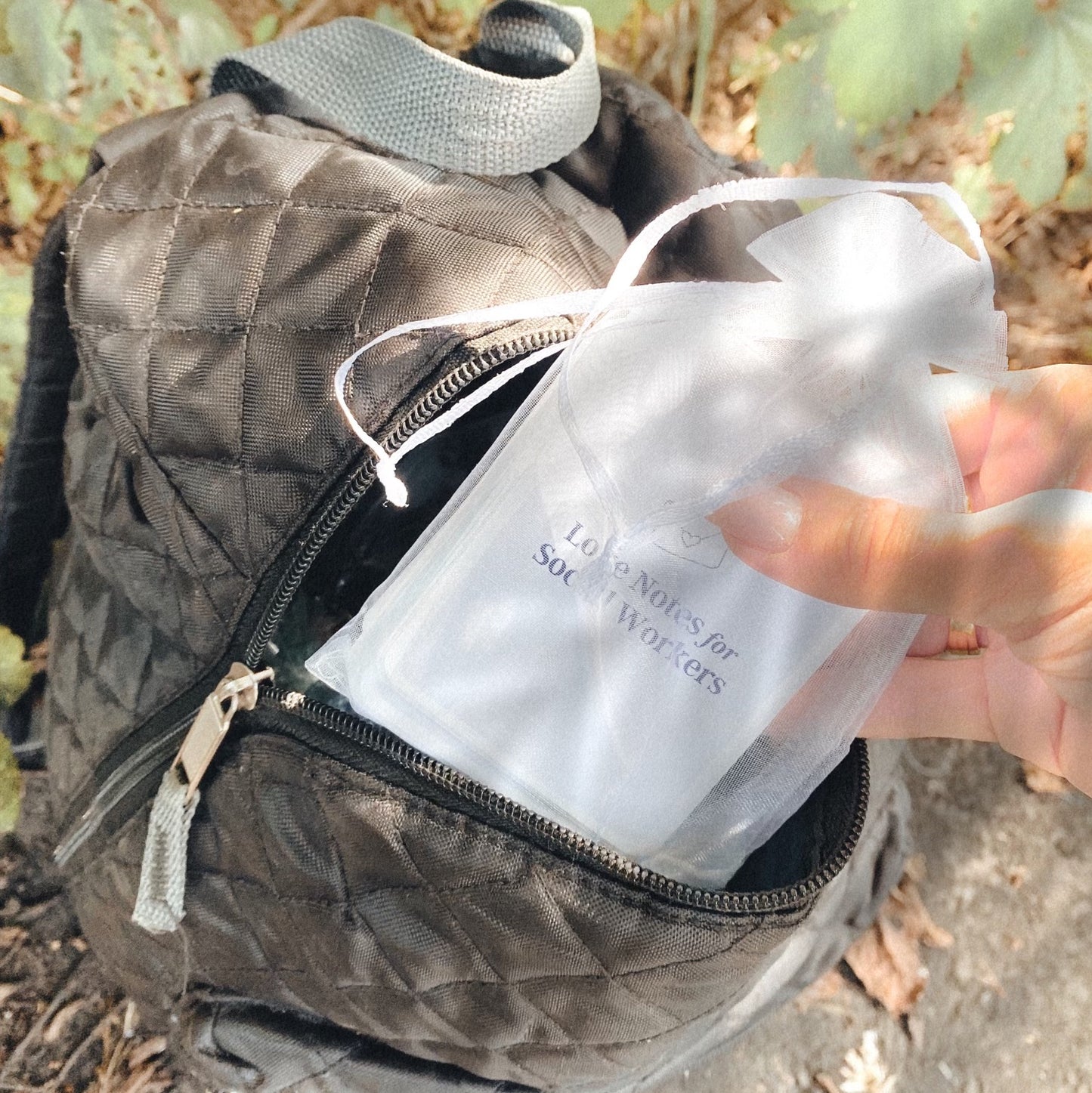 A woman's hand is pulling the card deck out of a black backpack. The card deck is inside a white organza bag and the top card reads "Love Notes for Social Workers".