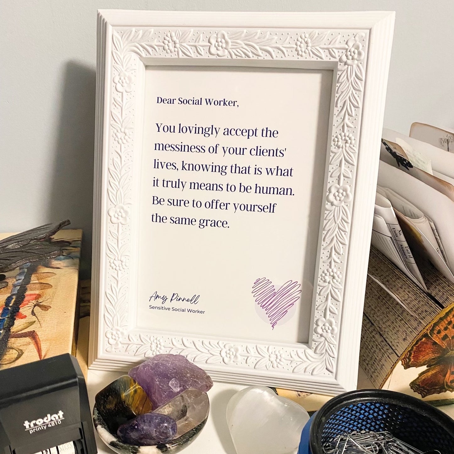The print is shown inside a white frame, on a white desk surrounded by office items. The print is white with dark purple font and reads "Dear Social Worker, You lovingly accept the messiness of your clients' lives, knowing that is what it truly means to be human. Be sure to offer yourself the same grace. Amy Pinnell, Sensitive Social Worker". In the bottom right corner of the print is a sketch of a scribbled purple heart.
