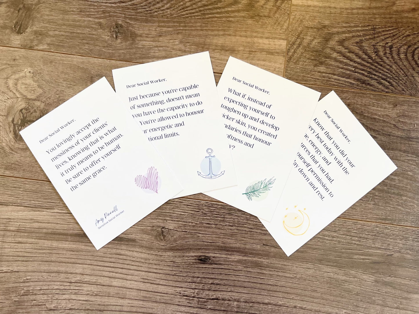 4 prints are fanned out on a wooden surface. Each print is white with dark purple font. Each print has a colorful sketch in the bottom right corner. The top card reads "Dear Social Worker, You lovingly accept the messiness of your clients' lives knowing that is what it truly means to be human. Be sure to offer yourself the same grace. Amy Pinnell, Sensitive Social Worker". The sketch in the bottom corner is a purple heart.