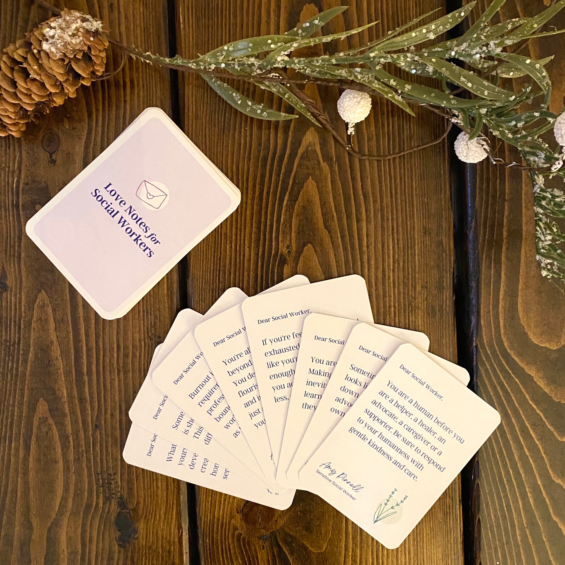 The card deck is stacked on a brown table beside a Christmas garland. The back  of the card deck is purple and says "Love Notes for Social Workers" on them. 8 cards are fanned out on the table and the top one reads:  "Dear Social Worker, You are a human before you are a helper, a healer, an advocate, a caregiver, or a supporter. Be sure to respond to your humanness with gentle kindness and care."