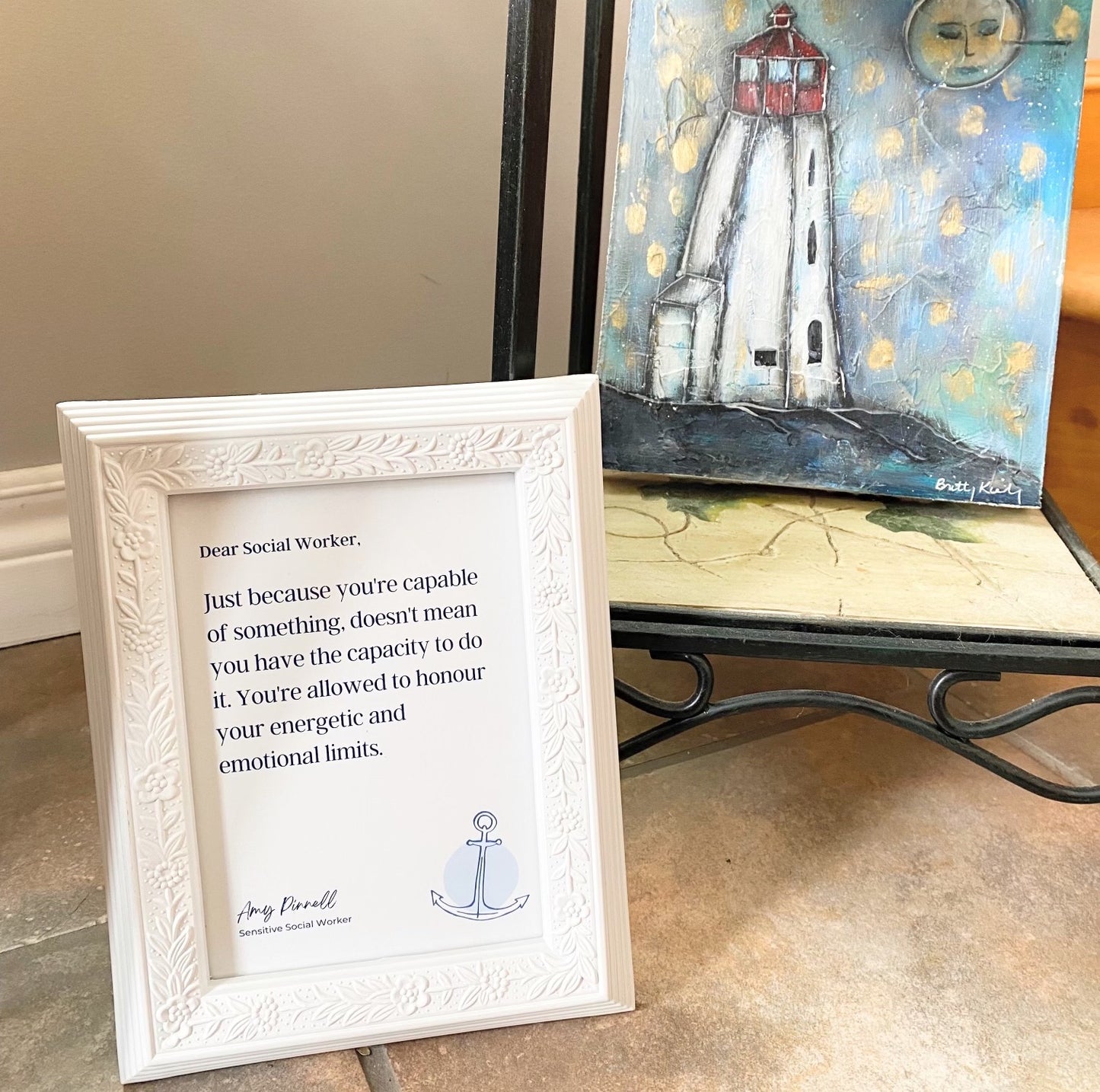 The print is shown in a white frame sitting beside a painting of a lighthouse. The print is white with dark purple font and reads "Dear Social Worker, Just because you're capable of something, doesn't mean you have the capacity to do it. You're allowed to honour your energetic and emotional limits. Amy Pinnell, Sensitive Social Worker". In the bottom right corner of the print is a sketch of a blue anchor.