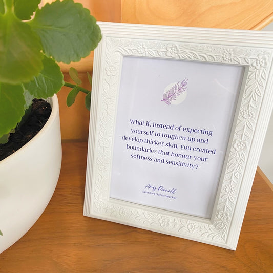 A light purple print with dark purple font is inside a white frame. The print reads "What if, instead of expecting yourself to toughen up and develop thicker skin, you created boundaries that honour your softness and sensitivity? - Amy Pinnell, Sensitive Social Worker". The frame is resting on a a wooden stand beside a green plant.