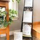 4 prints are displayed in white frames. 3 are shown a metal stand and one is one the floor. Beside the metal stand is a wooden stand filled with green plants. The prints are white with purple font with small colorful sketches in the bottom right corner.