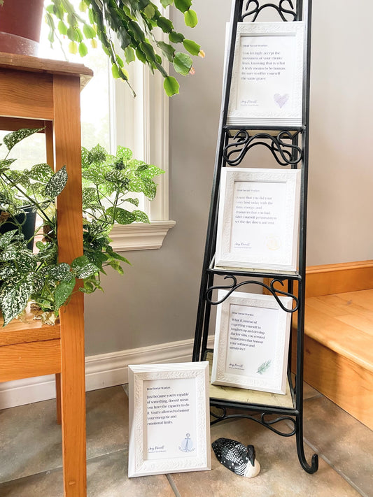 4 prints are displayed in white frames. 3 are shown a metal stand and one is one the floor. Beside the metal stand is a wooden stand filled with green plants. The prints are white with purple font with small colorful sketches in the bottom right corner.