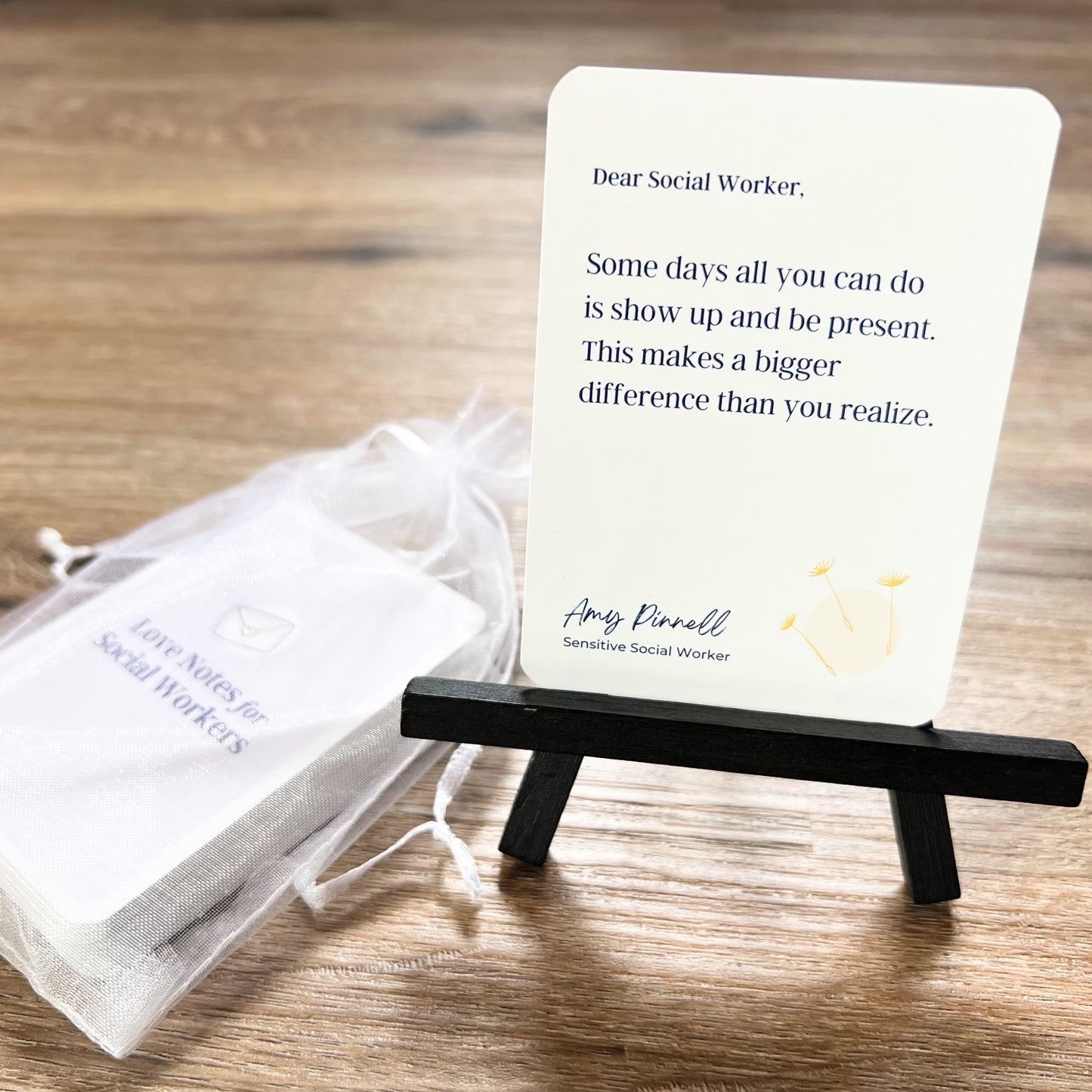 A single card is displayed on a small black stand. The card shows the message "Dear Social Worker, Some days all you can do is show up and be present. This makes a bigger difference than you realize". It is signed by Amy Pinnell, Sensitive Social Worker and includes a drawing of 3 yellow dandelion seeds. The rest of the card deck can be seen stacked inside a white organza bag behind the stand. The back of the card deck reads "Love Notes for Social Workers".  